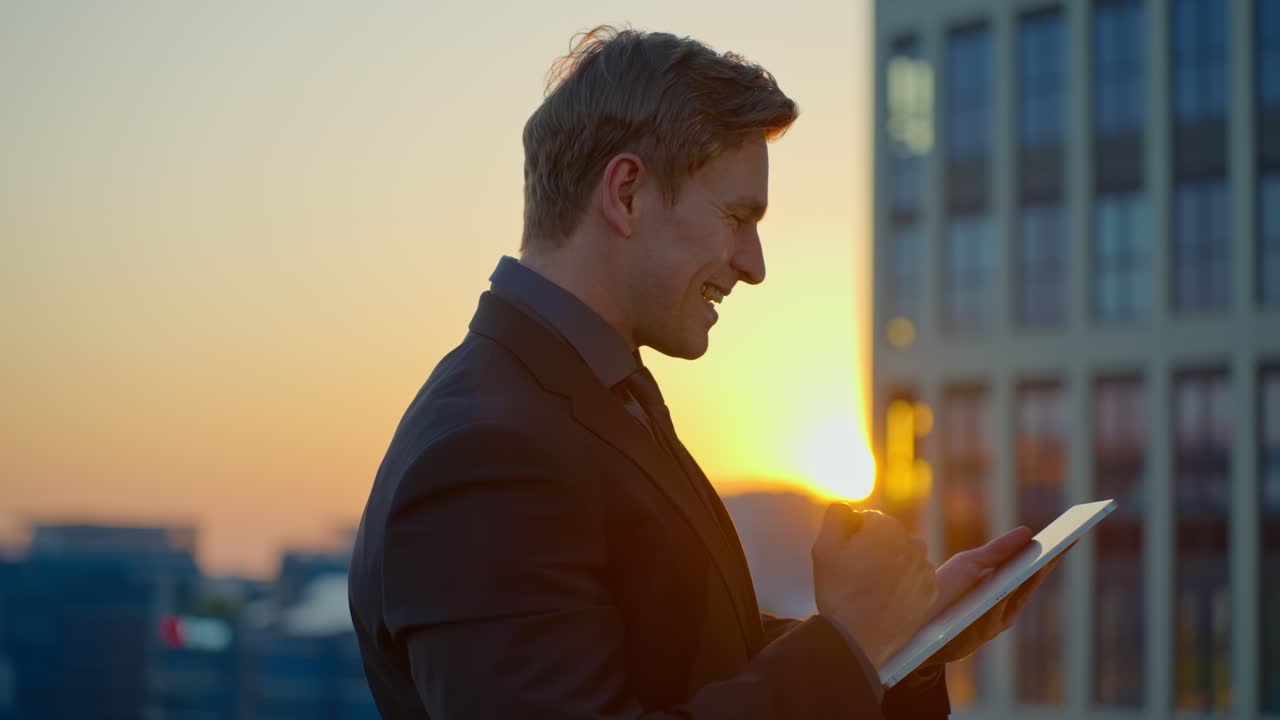 A happy Caucasian businessman in a suit celebrates a successful deal or profitable investment with a triumphant gesture while using a tablet on a modern office building rooftop at sunset