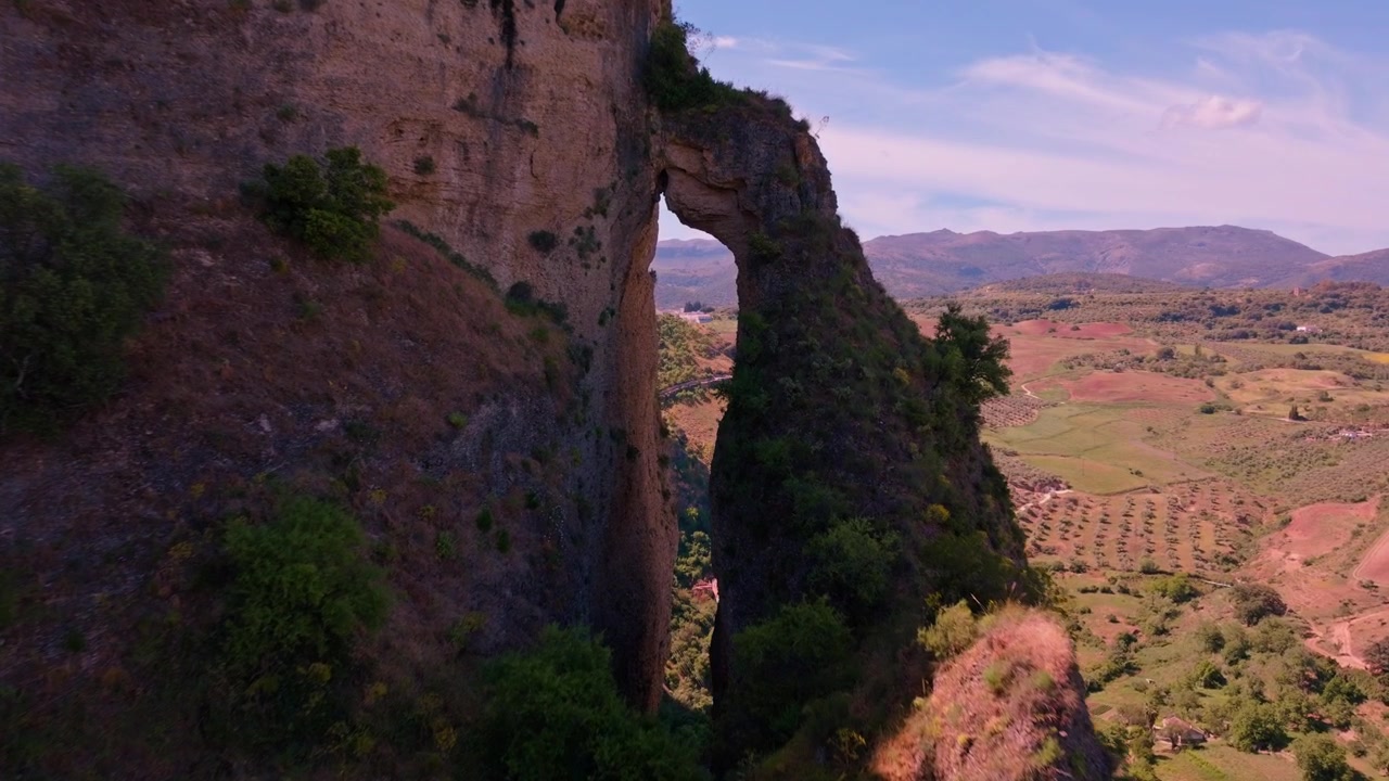 Riprese con drone che avanzano lentamente attraverso un teleobiettivo verso la cavità naturale scavata nella parete rocciosa di Ronda, con campi coltivati primaverili e cime distanti che attendono dall'altra parte
