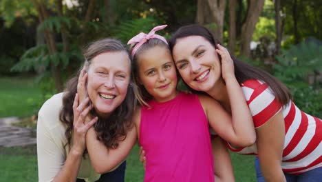 Portrait-of-happy-caucasian-grandmother,-granddaughter-and-mother-embracing-in-garden-and-smiling