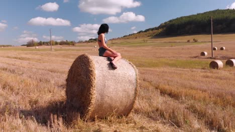 Toma-Aérea-De-Una-Hermosa-Morena-Tomando-El-Sol-Mientras-Se-Sienta-En-Un-Fardo-De-Heno