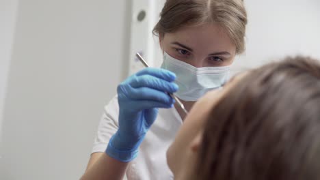 young female dentist in mask approaches with tools. dental lamp lights into a patient's mouth. standing upon a patient