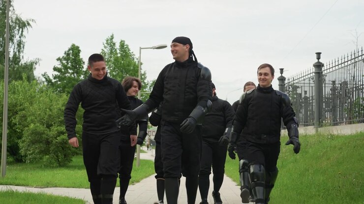 Gruppo di uomini con attrezzatura protettiva nera che camminano lungo un sentiero lastricato con erba verde e alberi su un lato, una recinzione metallica oltre. Le calzature includono scarpe da ginnastica o stivali, alcuni indossano ginocchiere mentre altri