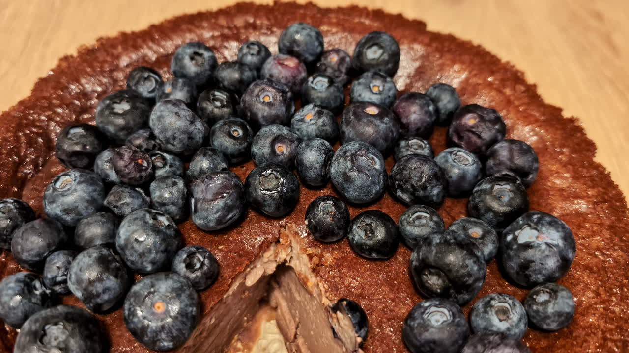 A zoomed-out view of a chocolate cake topped with blueberries, showing a cut slice and creamy interior