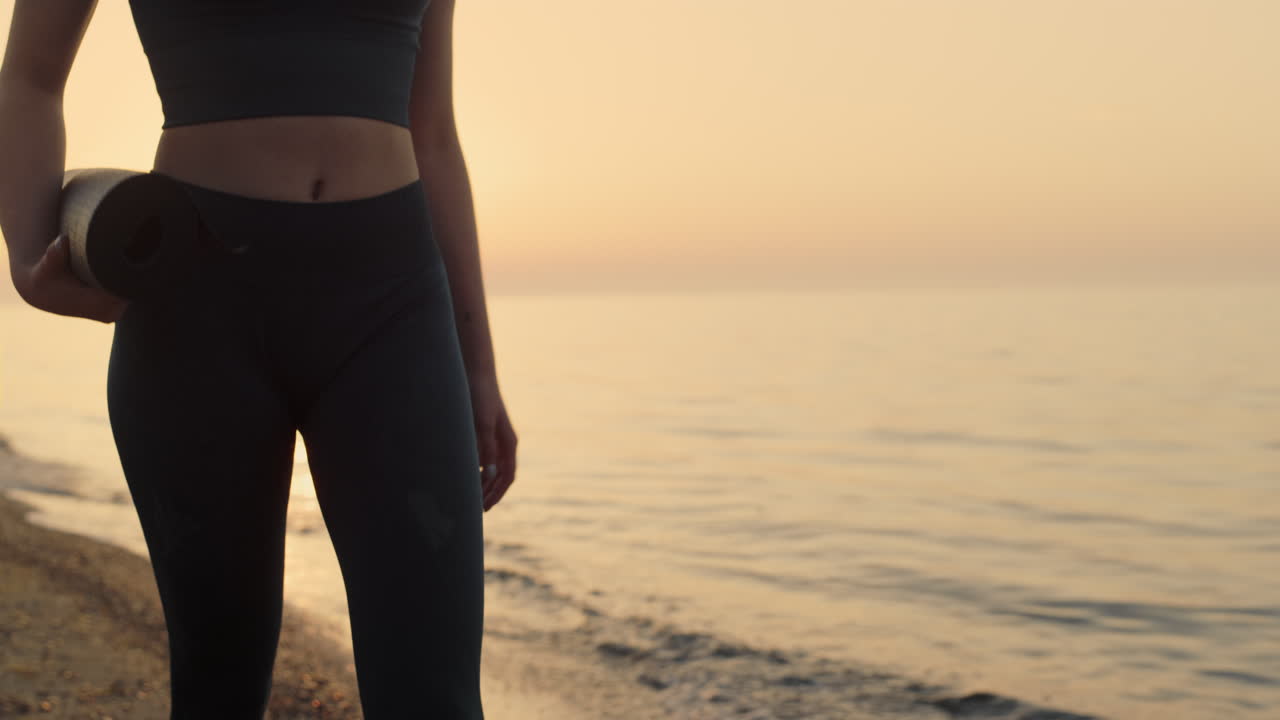 Fit girl holding mat walking seashore at sunset. Sportswoman going to training.