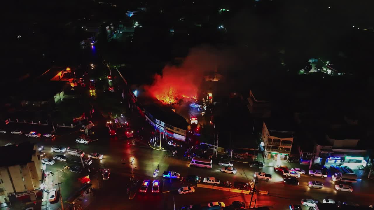 Wide establishing shot of a major building fire at night, showing large scale emergency response with many vehicles and personnel, Santiago de Los Caballeros, Dominican Republic. Aerial