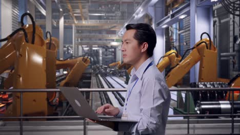 side view of an asian male professional worker standing with industrial robotic arms working, observes by looking up before he come to concentrating on the laptop and keep on typing