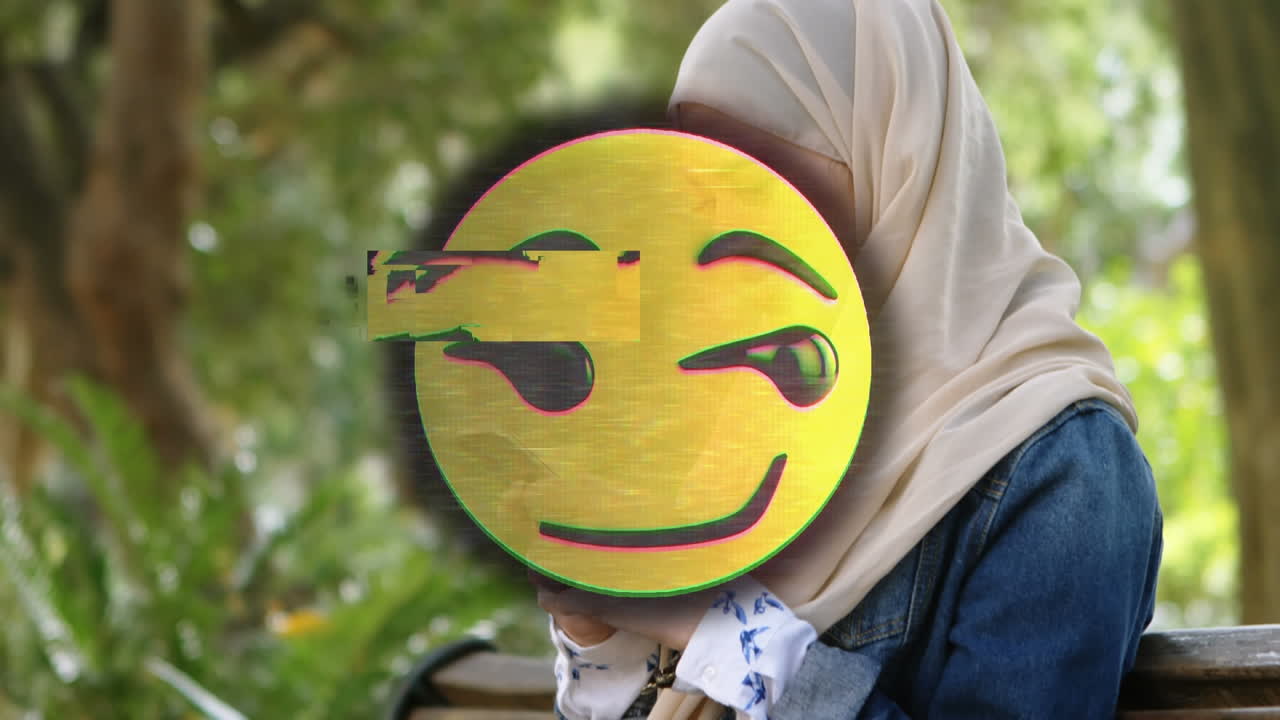 Animation of happy emoji over woman in hijab using smartphone