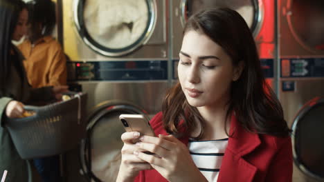 pretty stylish woman tapping and texting message on smarphone while sitting in laundry service room