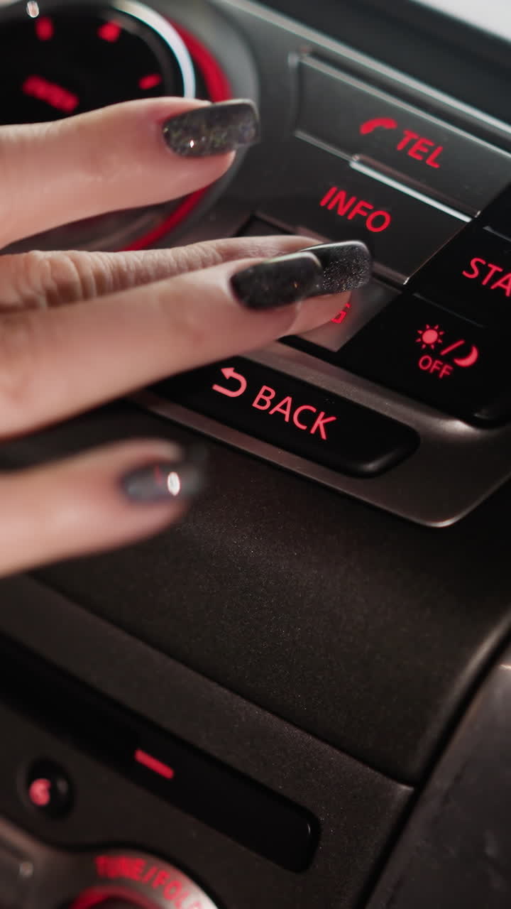 First-person view of a woman's hand pressing the settings button on a car control panel at night. The dashboard is illuminated with red lights, highlighting the controls. Shot with a handheld camera