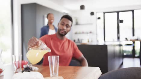Glückliches-Afroamerikanisches-Paar,-Das-Saft-Einschenkt-Und-Frühstück-In-Der-Sonnigen-Küche-Serviert,-Zeitlupe