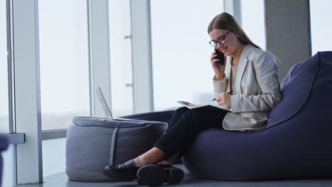 young business lady sits in chair and talks on the phone. left-handed woman making notes in her paper notebook. panoramic windows in blur at backdrop.