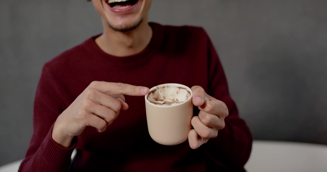 Uomo adulto africano che sorride, solleva occhiali con la scritta "MERRY" e sorseggia cioccolata calda per festeggiare in studio, con un maglione.
