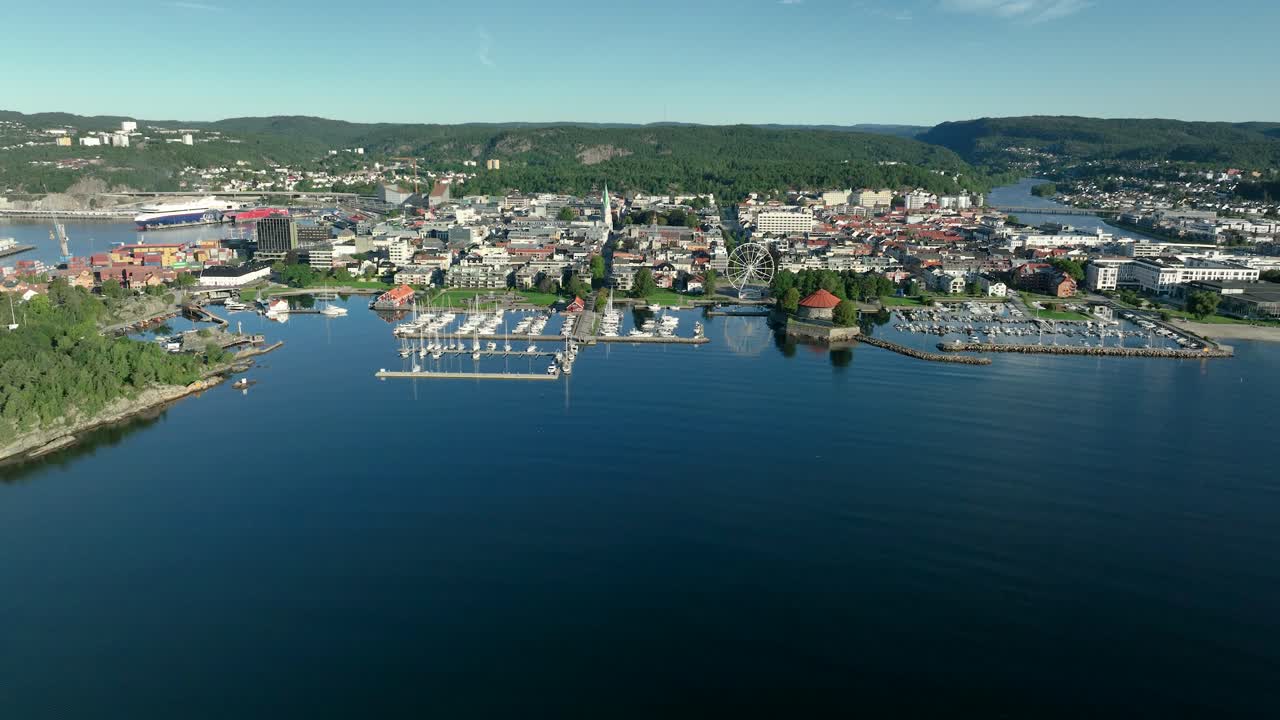 This aerial view captures the vibrant waterfront of Kristiansand in Norway featuring a marina filled with boats, a ferris wheel, and lush hills in the background under a clear blue sky