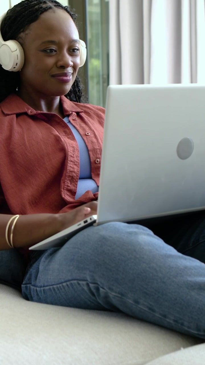 Vertical video: Shifting laptop angle, woman in rust shirt typing on sofa working with headset