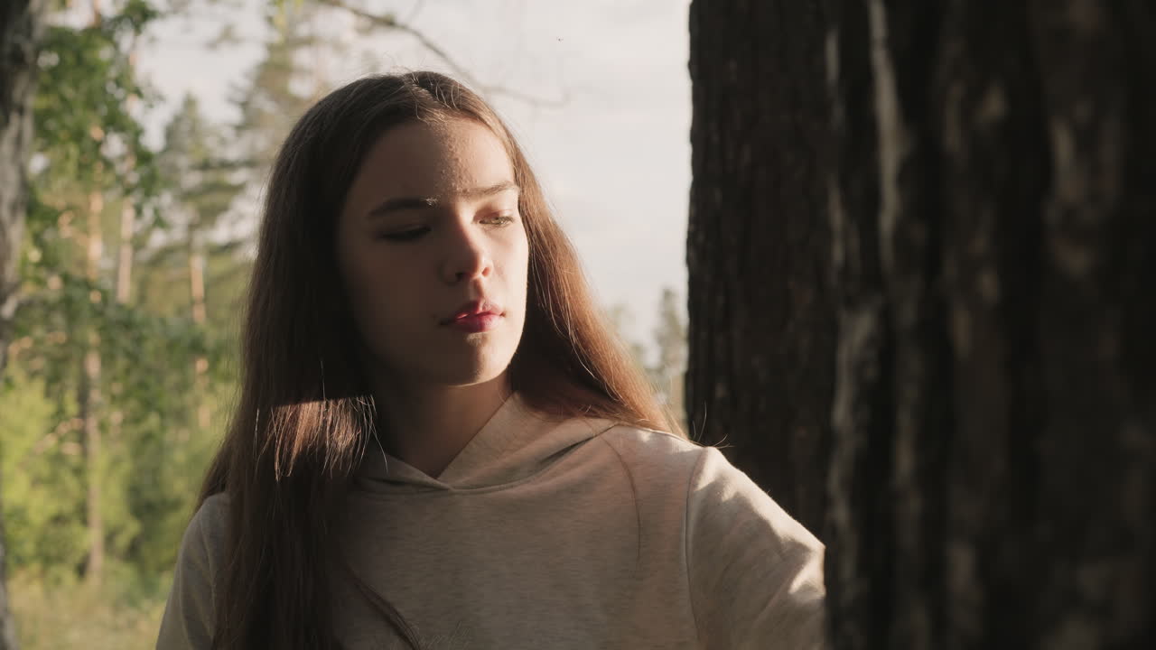 슬픈 여자가 해가 지는 빛으로 비추는 나무 근처에 서 있습니다. 긴 느슨한 머리카락을 가진 젊은 여자는 숲에서 과거의 관계에 대해 생각합니다. 스트레스를 해소하기 위해 자연에서 취미