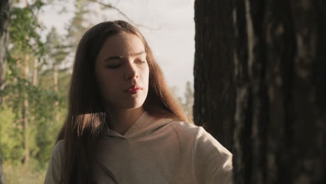 sad woman stands near tree lit by setting sun light. young lady with long loose hair thinks about past relationship in forest. pastime in nature to relieve stress