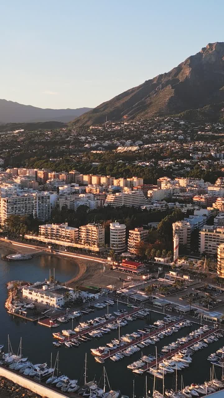 Vista aerea verticale del porto di Marbella al tramonto dorato, che mostra le barche del porticciolo, le calme acque del Mediterraneo e le montagne sullo sfondo