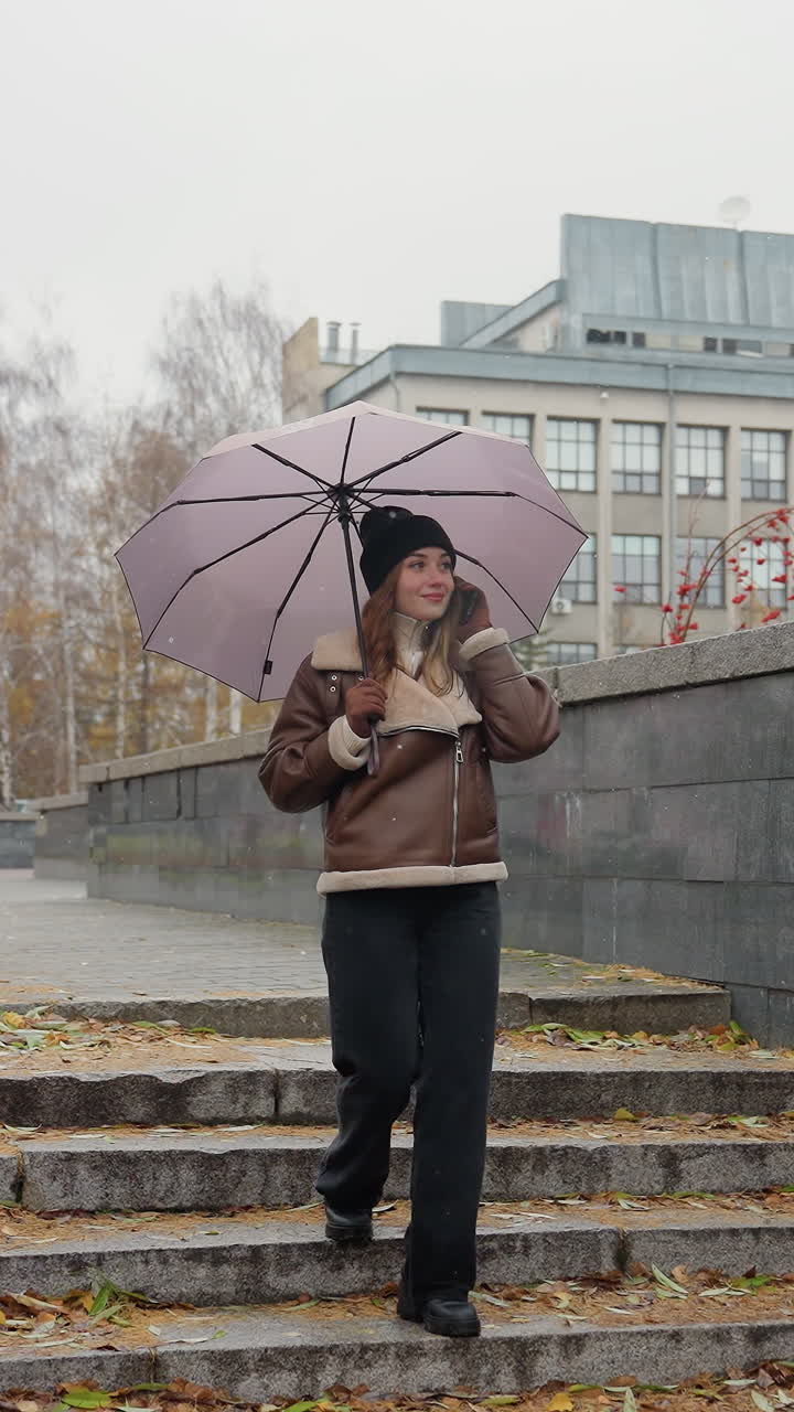 Studente sorridente al telefono che tiene un ombrello, indossa un berretto di lana, una giacca in shearling marrone, pantaloni neri, mentre scende dei gradini in pietra durante una leggera nevicata con foglie autunnali colorate