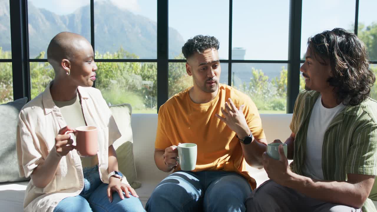 African American friends sitting on sofa holding mugs near window center man glancing sharing point