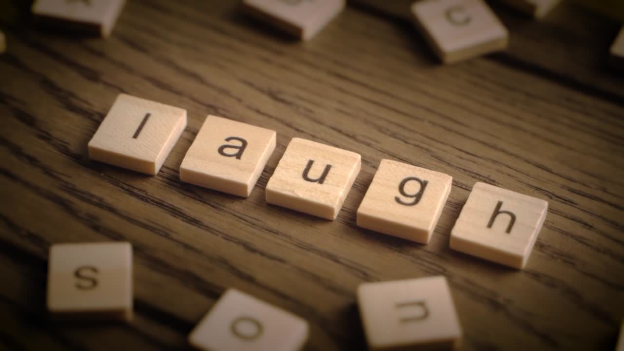 Wooden scrabble-style tiles spell out laugh as the camera pans over a rustic wood surface in warm lighting.