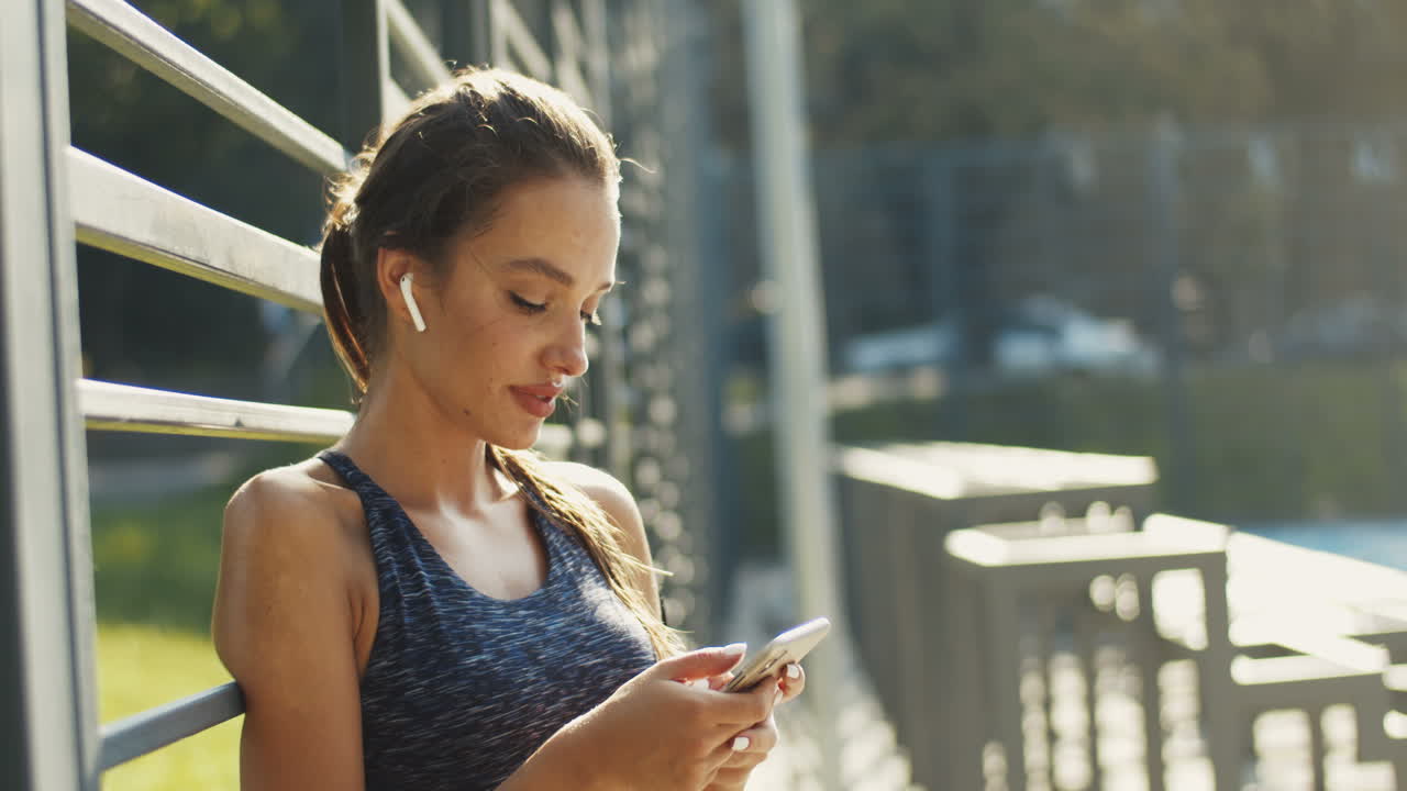 chica deportiva con airpods riendo mientras envía mensajes de texto en el teléfono inteligente después de hacer ejercicio en la cancha al aire libre en un día de verano