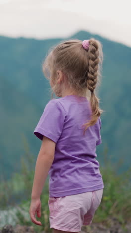 cute little girl with braided hair looks at old distant mountain foggy silhouettes and scratches leg at highland backside view slow motion