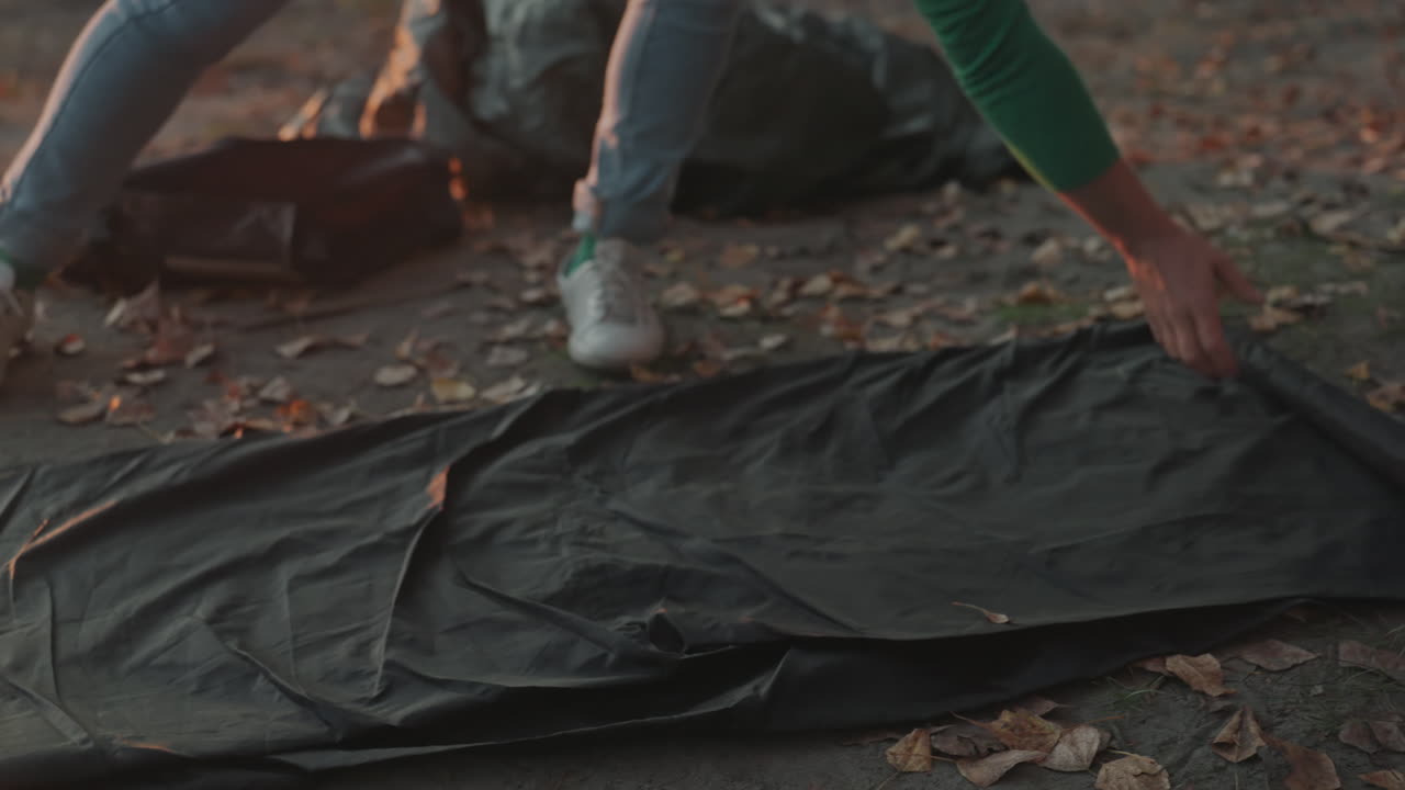 Un campeggiatore srotola un sacco a pelo su un marciapiede cosparso di foglie autunnali, con le mani che lisano il tessuto e raddrizzano le cuciture, jeans e scarpe da ginnastica visibili nella calda luce del crepuscolo, mentre prepara il bivacco al campeggio.