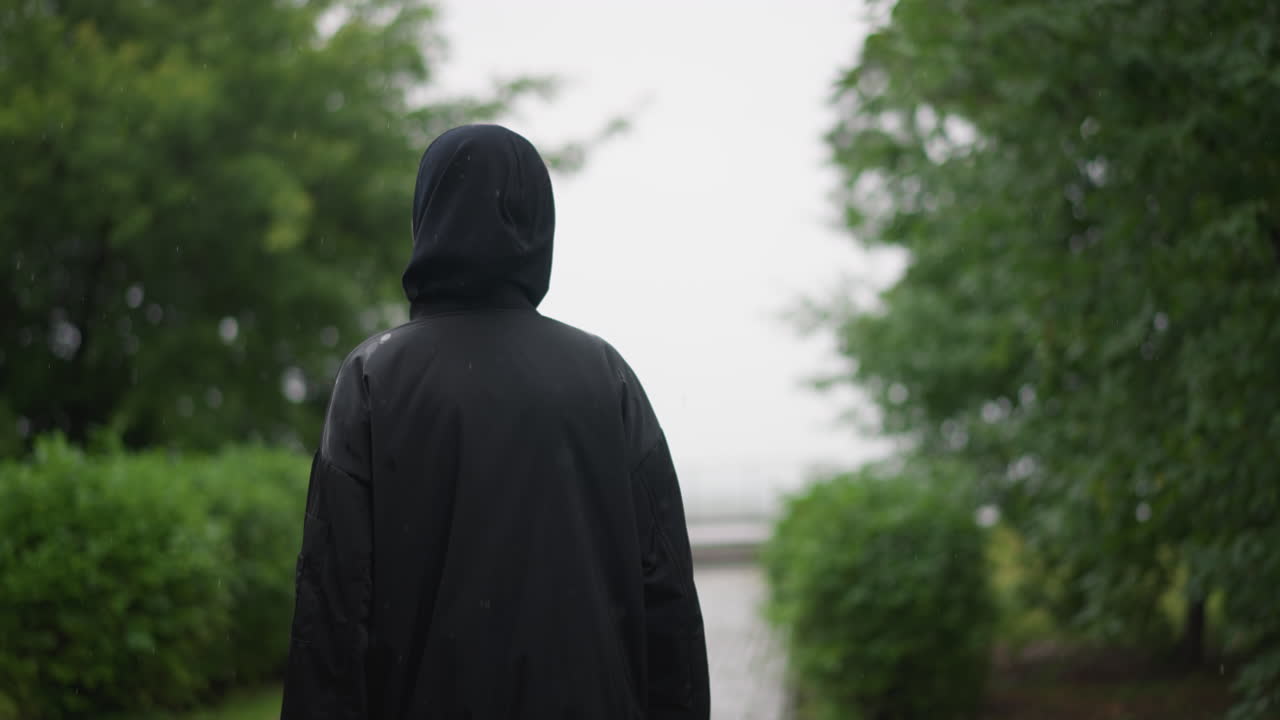 Quiet female figure close to water droplets. Woman wearing hood next to glistening puddles beneath rain. Female figure protected from rain with hood near sparkling water pools under cloudy sky