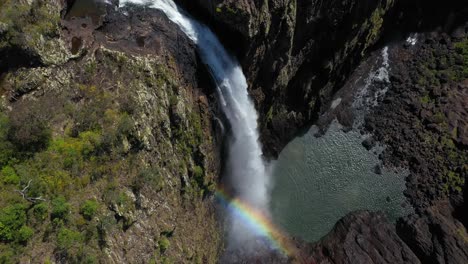 Waterfall-cascading-over-canyon-rock-cliff,-rainbow-mist,-aerial-zoom