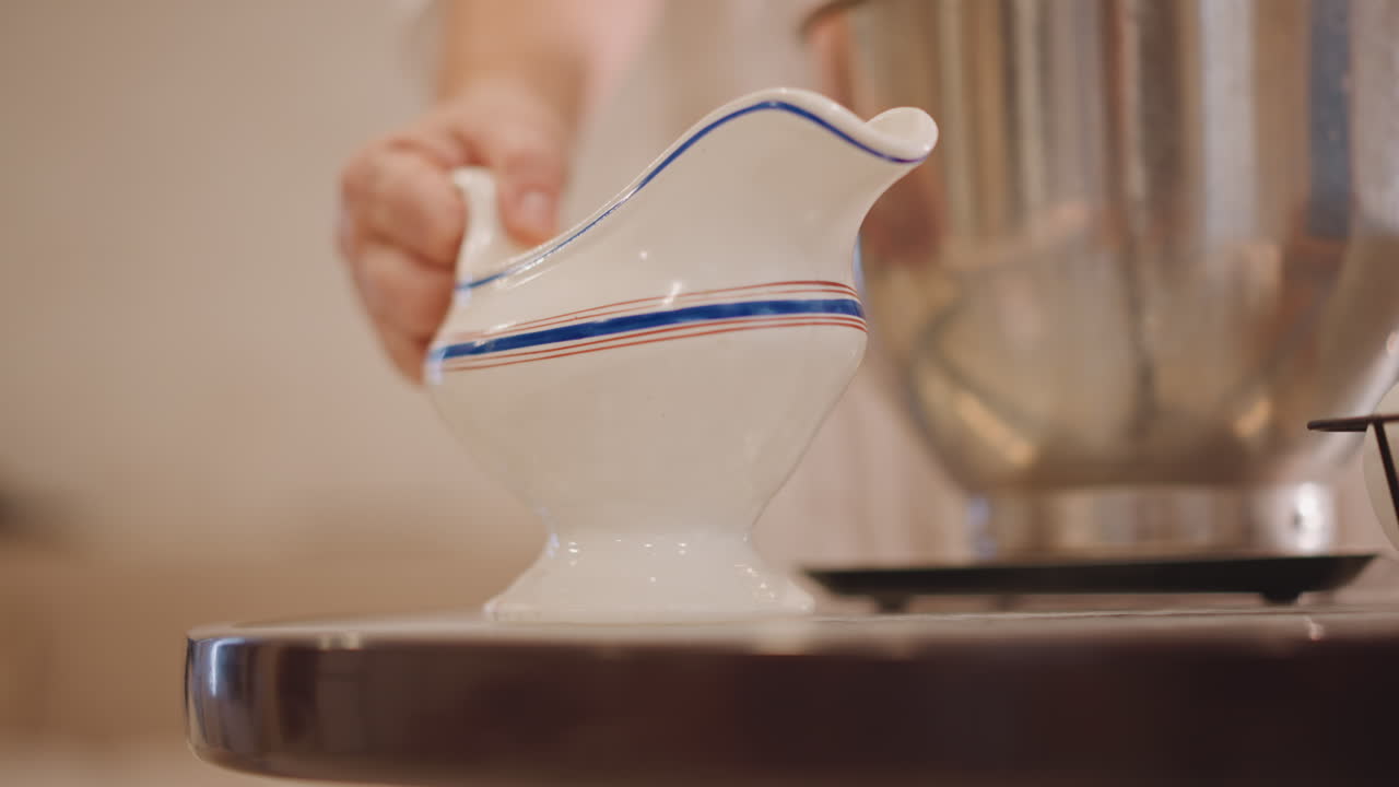 Mano che abbassa una ciotola mescolatrice in acciaio inossidabile. Un bagliore caldo e delicato illumina una salsiera in porcellana e una ciotola mescolatrice. Primo piano dettagliato di una salsiera in porcellana con una mano e una ciotola mescolatrice in un'accogliente cucina.