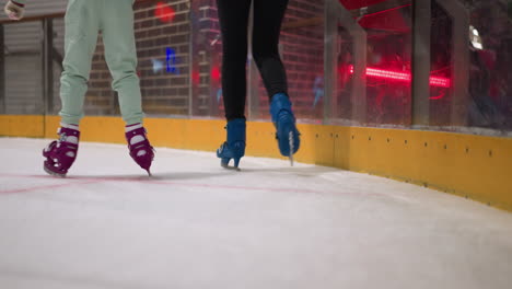 close view of two people skating on an ice rink, one in purple skates and mint green pants, the other in blue skates and black pants, against a backdrop of red lights and a brick wall