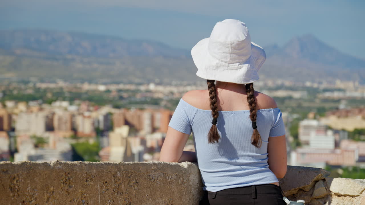 Una ragazza viaggiatrice con un cappello da pescatore bianco ammira la costa da un belvedere ventilato in cima a una collina ad Alicante, Spagna