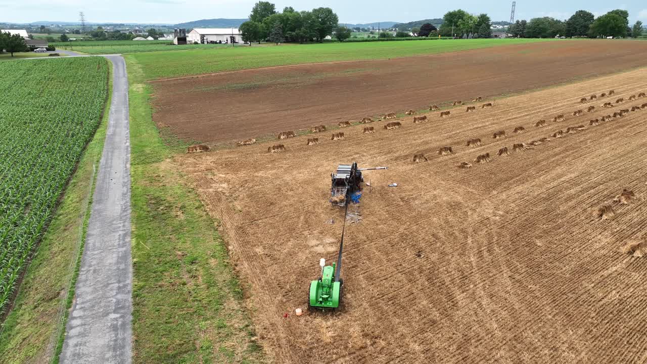 An aerial circling view of a threshing machine and tractor in a wheat field that is being harvested in a historic way
