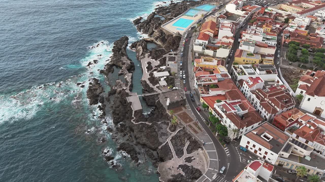 Bird's-eye view of Tenerife coastline showing the waves crashing on the rocks, the pools and the iconic colorful beach houses and the busy road in between the coasline and the city