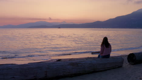 una mujer joven se sienta y contempla contemplativamente la hermosa puesta de sol en una playa tranquila con vistas a las montañas en la hermosa columbia británica, canadá