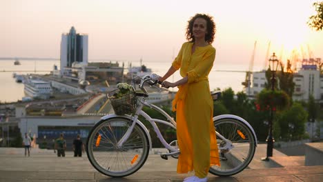 young brunette woman in long yellow dress standing by her city bike holding its handlebar with flowers in its basket during the