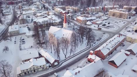 Vehicles-Driving-Between-Buildings-And-Smiltene-Evangelical-Lutheran-Church-Covered-With-Snow-At-Winter-Season-In-Latvia