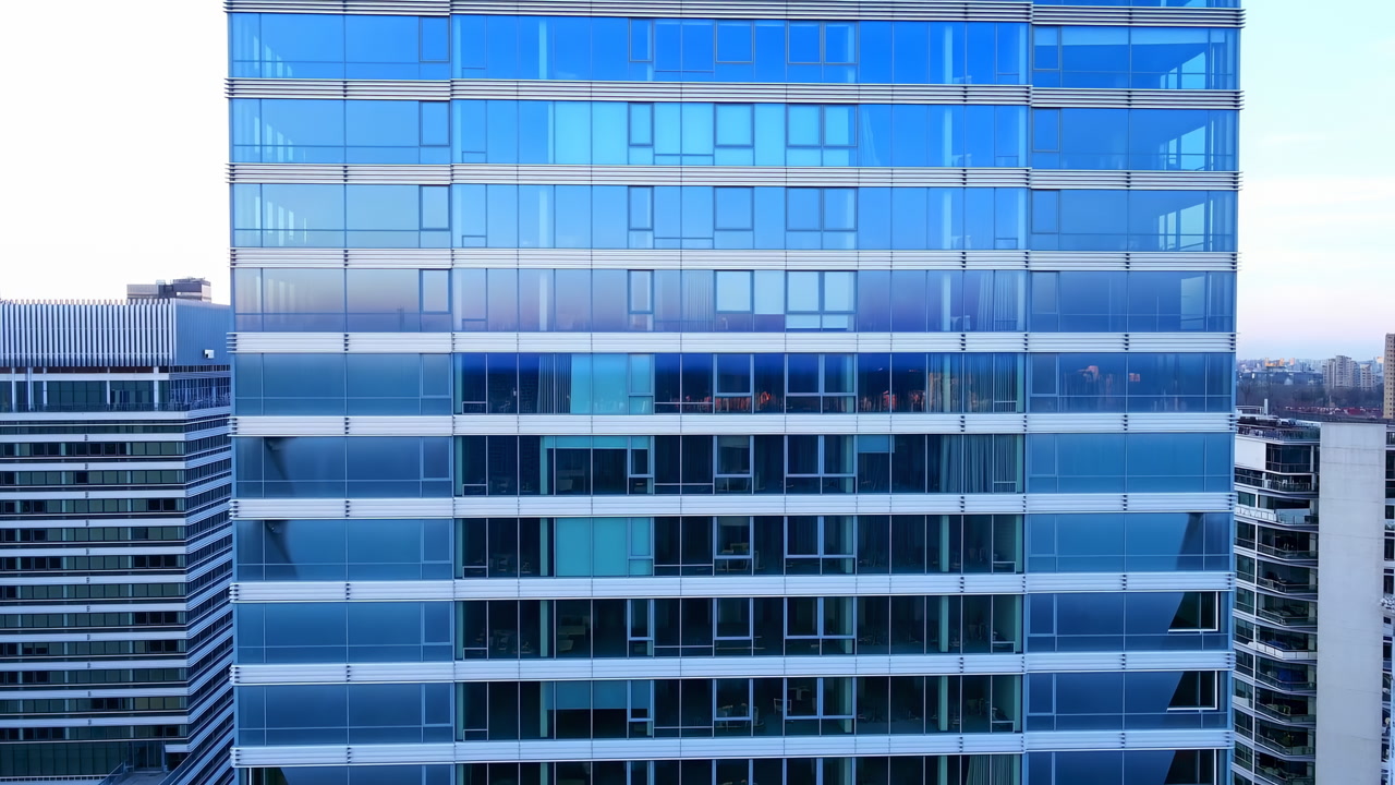 Blue Glass Office Tower Facade Showcasing Mirrored Skyline And Gridlike Windows During Aerial Drone Sweep