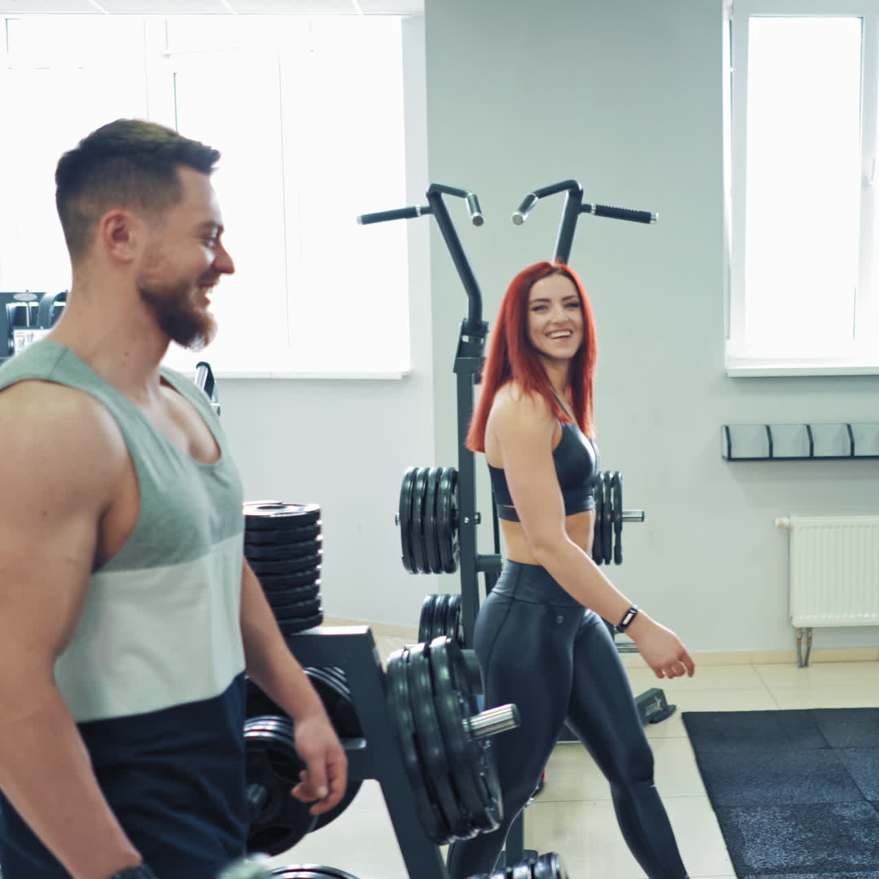Young attractive woman talking to a strong male trainer at the gym. Happy athletic couple having a talk while coming into the sports center on the background of modern simulators.