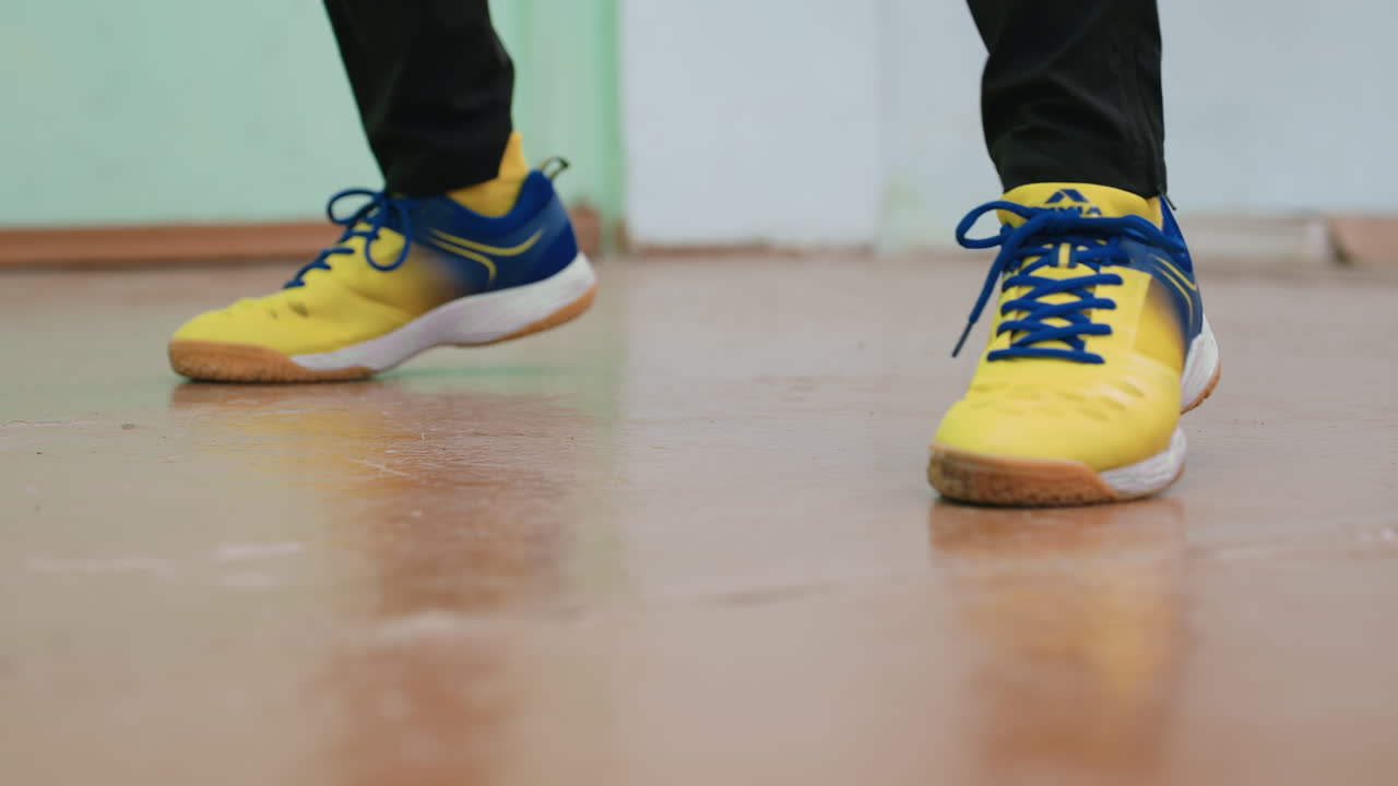 Lower angle close up of athlete wearing yellow blue sports shoes practicing footwork indoors, showing active steps, agility, and movement on polished floor with energy during training session