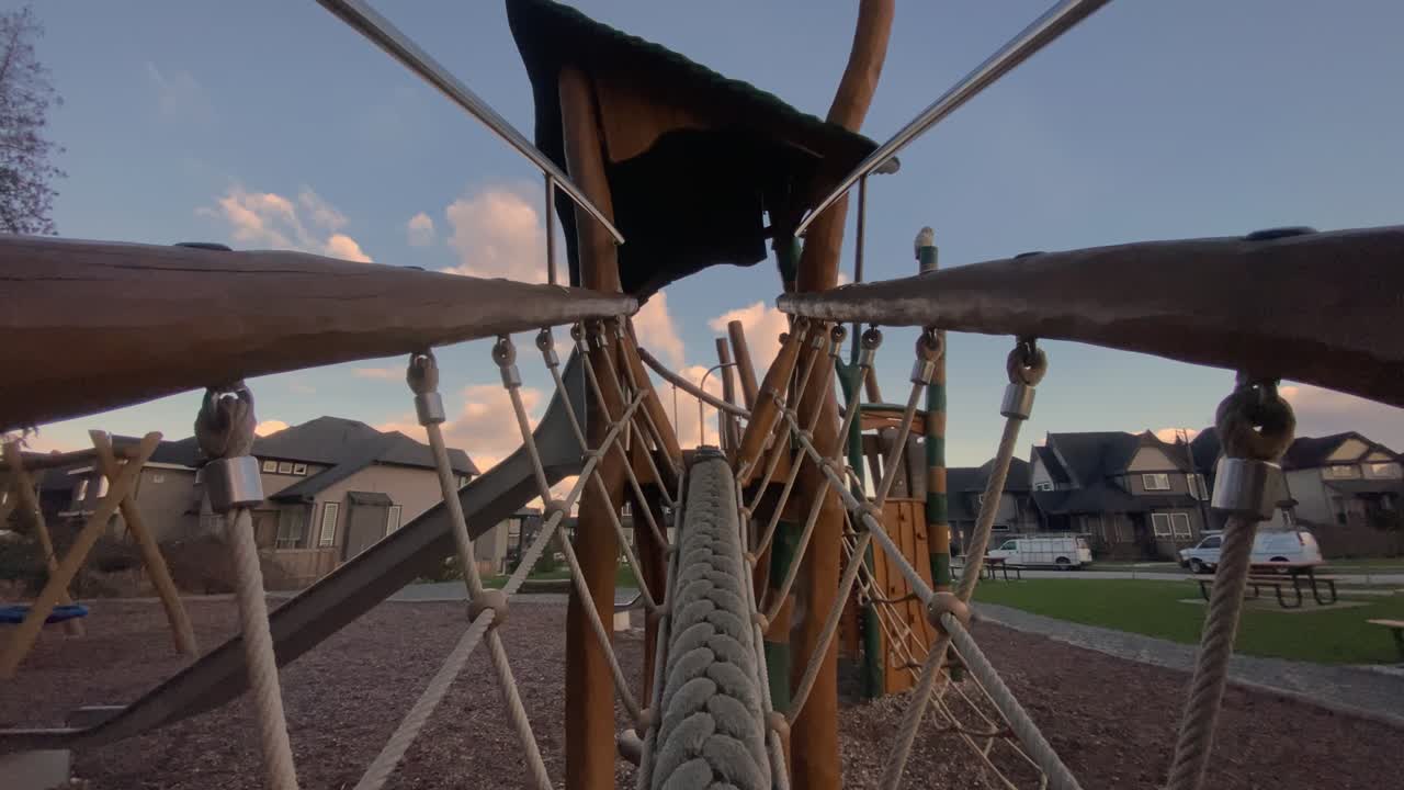 Thick woven climbing rope stretching across timber play structure hanging above wood chips framed against suburban housing beneath cloudy sunset sky captured via backward moving viewpoint