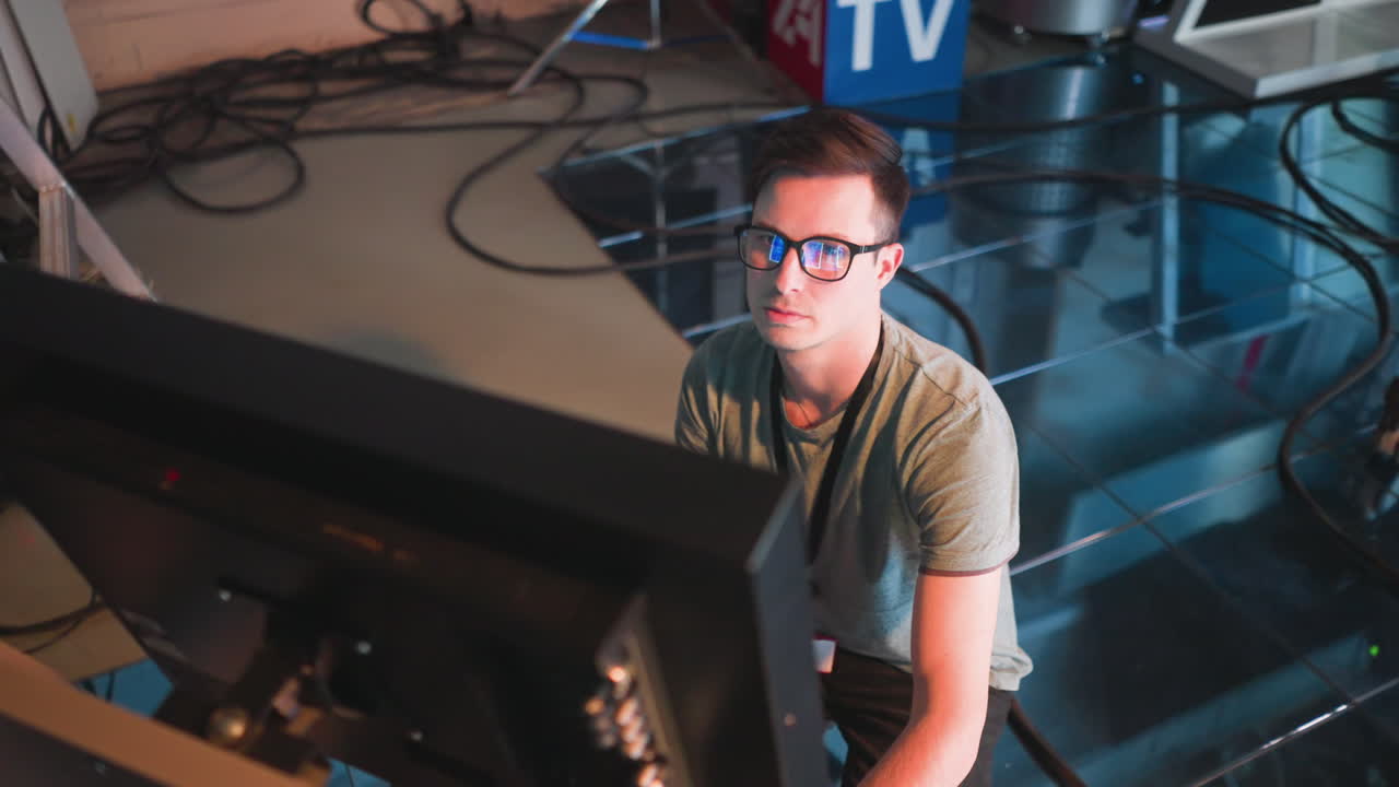 Person working in TV studio, wearing glasses, focused on computer monitor. Close-up shot showing intense concentration, modern tech environment, cables, and lighting setup in background