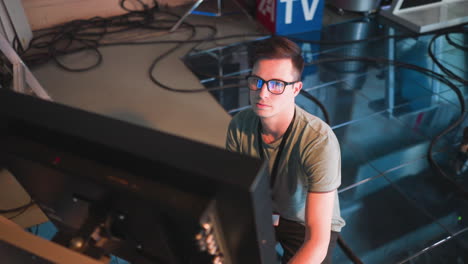 person working in tv studio, wearing glasses, focused on computer monitor. close-up shot showing intense concentration, modern tech environment, cables, and lighting setup in background