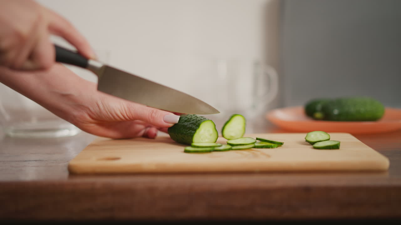 Close up hand view of fair skinned person carefully slicing fresh cucumber on wooden cutting board in kitchen with other utensils and glassware placed neatly on modern countertop
