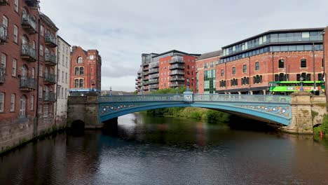 a green double-decker bus travels across a blue bridge spanning the river aire, surrounded by victorian brick buildings under daylight with steady camera movement