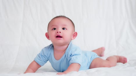 happy family, cute asian newborn baby wear blue shirt lying, crawling play on bed looking at camera with laugh smile happy face. innocent little new infant adorable. parenthood and mother day concept.