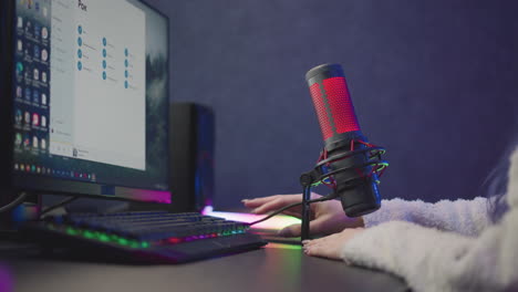 lady in fluffy jacket reaches forward to reposition red lit studio microphone and cable next to rgb strip illuminated monitor keyboard setup before livestream recording session in gaming workspace