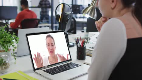mujer de negocios caucásica sentada en un escritorio usando una computadora portátil haciendo una videollamada con una colega
