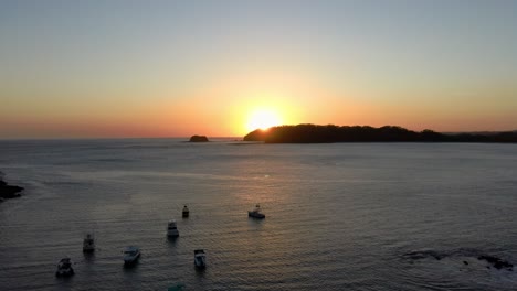 Hermosa-Puesta-De-Sol-Detrás-De-La-Isla-En-El-Horizonte-Con-Barcos-En-Una-Bahía-Tranquila,-Drones-4k