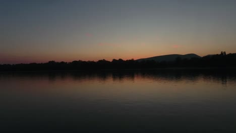 Toma-Aérea-De-Una-Puesta-De-Sol-O-Un-Amanecer-Sobre-El-Agua,-Que-Ascendiendo,-Elevándose-Para-Revelar-Una-Escena-Más-Amplia